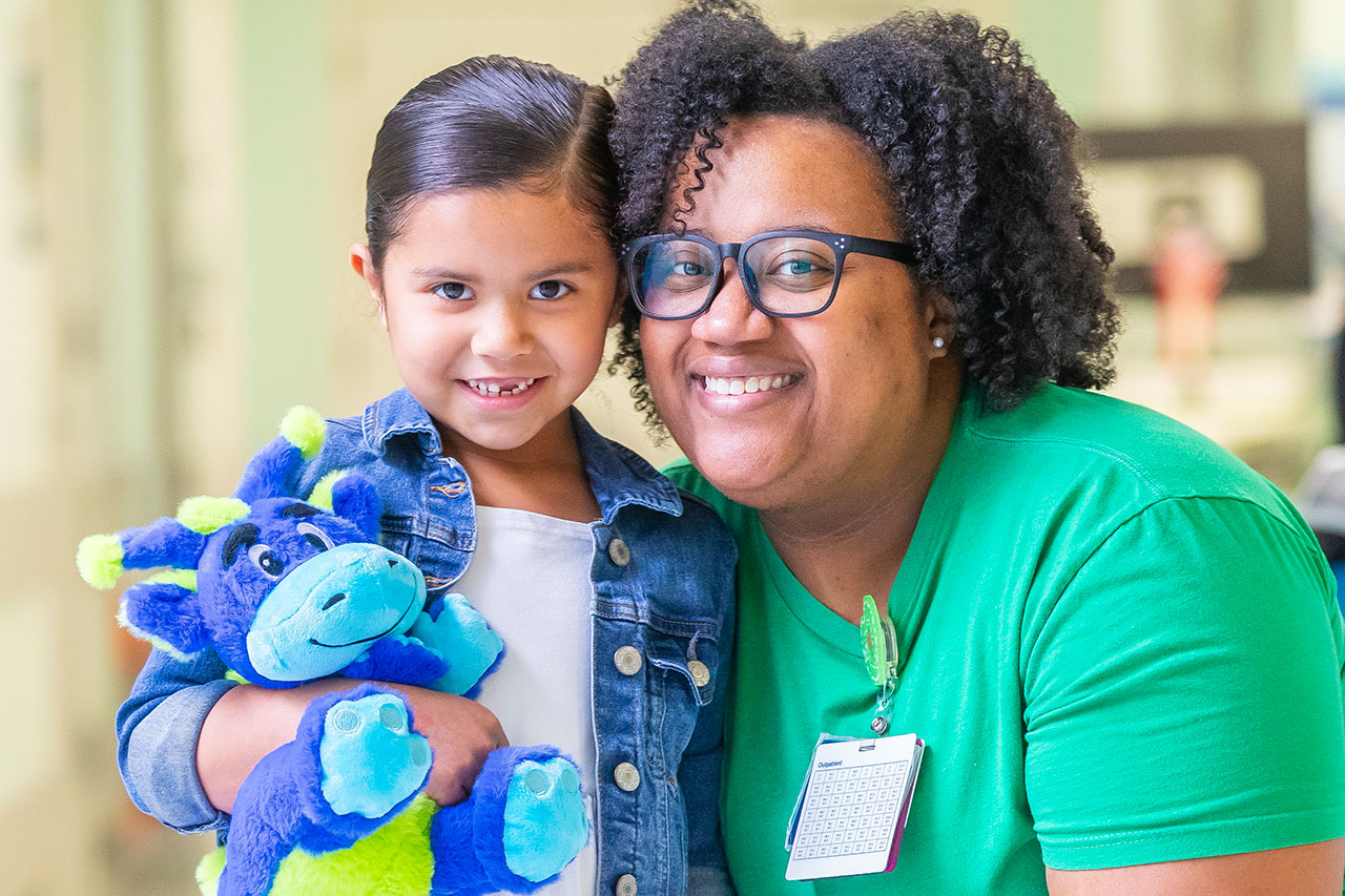 Smiling woman in glasses and girl holding a blue dragon stuffed animal. Woman wears an Outpatient ID