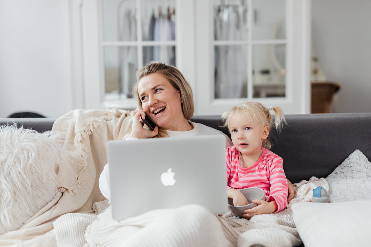 Laughing woman talks on phone with laptop; young child in pink stripes sits beside her, holding a bowl